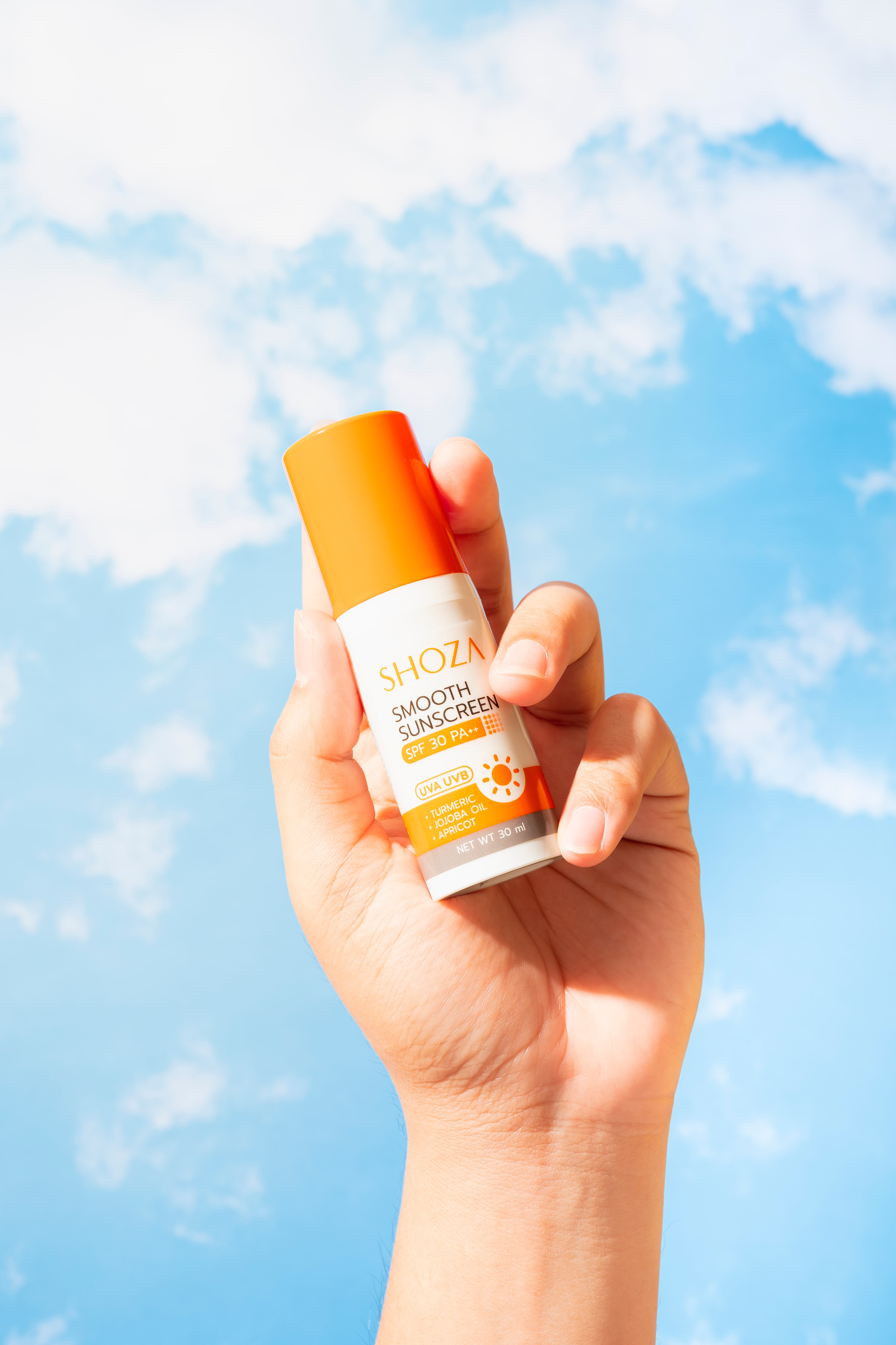 มือถือขวดกันแดด SHOZA Smooth Sunscreen ฝาสีส้ม ชูขึ้นบนพื้นหลังท้องฟ้าสีฟ้ามีเมฆ